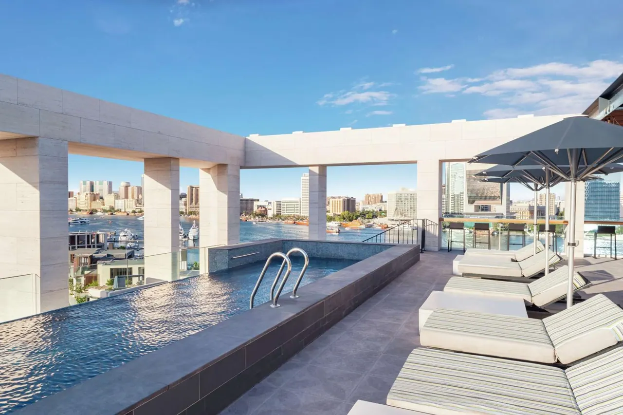 A rooftop pool with pool steps and sun loungers under grey shade umbrellas overlooks a waterway and city buildings under a blue sky.