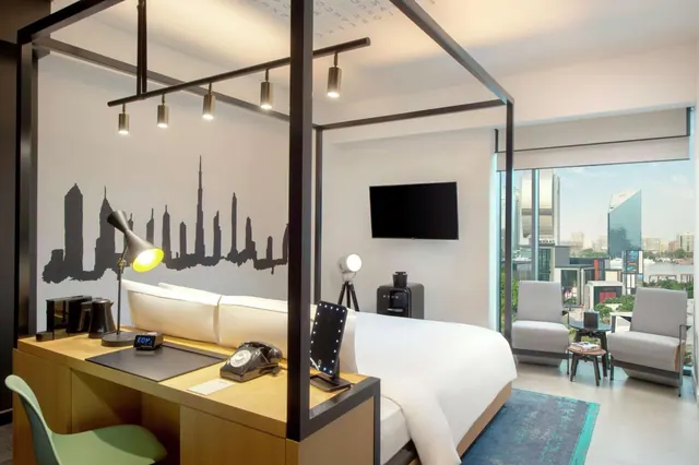 A hotel bedroom with a canopy-framed bed, a wall mural of a city skyline, a desk with a lamp and telephone, two armchairs, a wall-mounted TV, and floor-to-ceiling windows showing buildings outside.