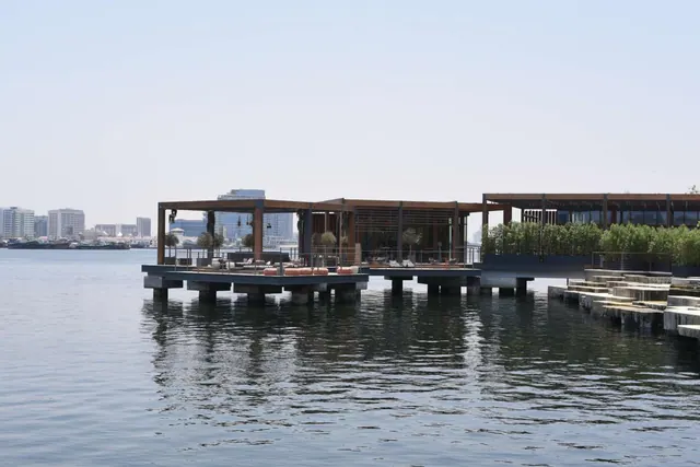 Wooden-roofed waterfront pavilions and a stepped terrace stand on stilts over calm water, with a city skyline across the water to the left.