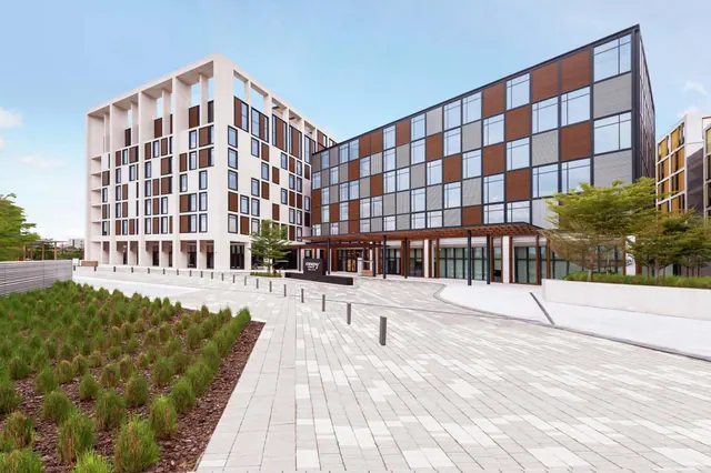 The exterior of a modern hotel building with a paved forecourt, an entrance canopy, and a facade of panelled sections and windows, with young trees and planted beds in front.