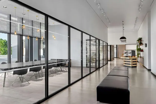A long corridor with glass walls, a meeting room with tables and chairs on the left, a black upholstered bench in the foreground, and a coffee machine counter at the far end.