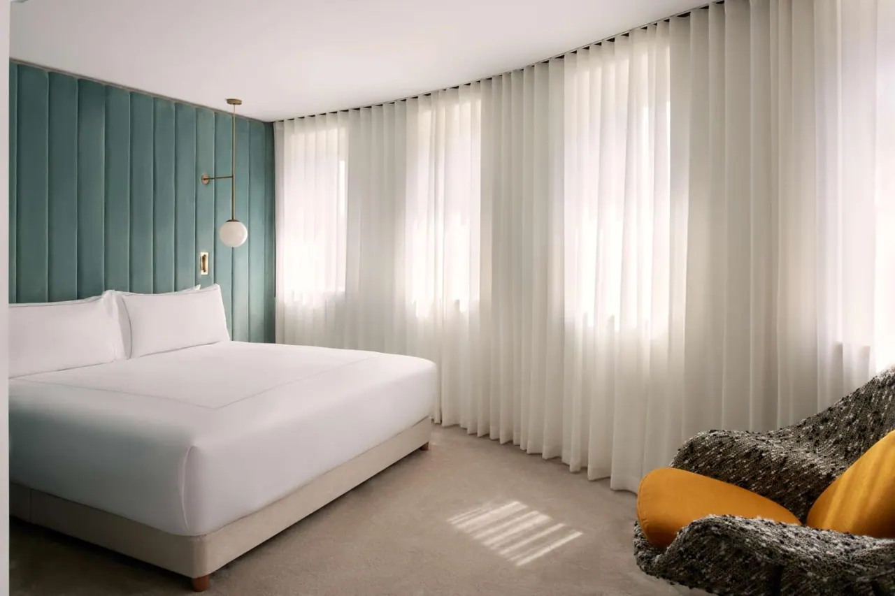 A bedroom with a made king bed, a teal upholstered headboard wall, sheer floor-length curtains and a tweed armchair with mustard cushions.
