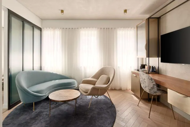 A living area with a curved blue sofa, a beige armchair and a round table on a dark rug, beside a desk with a wall-mounted television.