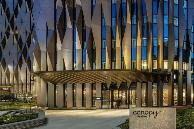 Exterior of a hotel building with an angular bronze-clad facade, a glazed entrance beneath a sloping canopy, and a stone sign reading canopy by Hilton, photographed in the evening.