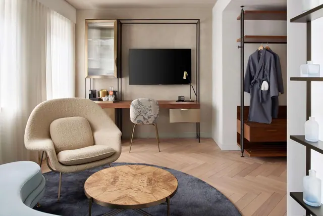 A hotel room with an armchair and round coffee table on a rug, a wall-mounted television above a desk, and an open wardrobe holding robes.