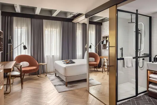 A hotel bedroom with a made bed, two armchairs and a desk, with a glass-walled walk-in shower visible on the right.