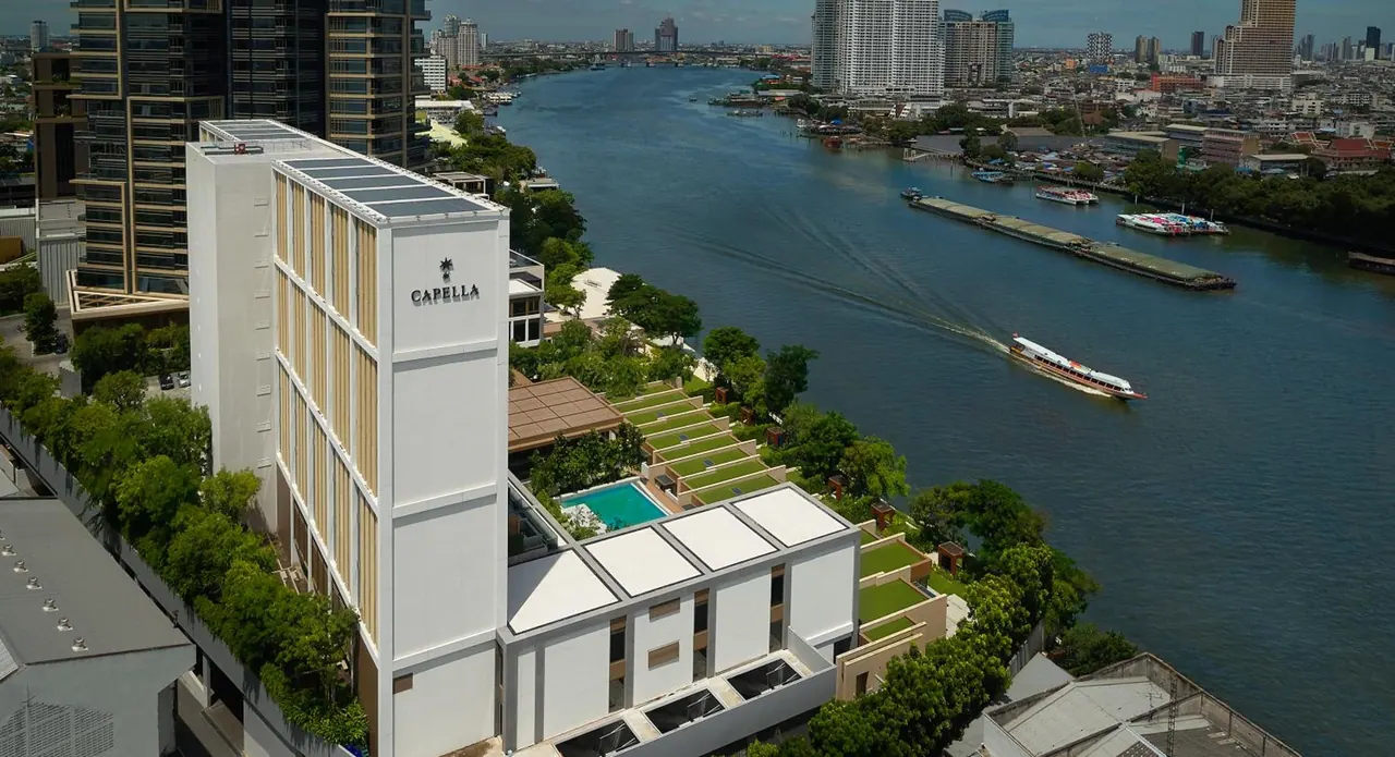 Aerial view of a white hotel building beside a wide river, with a small pool in the garden and boats on the water.