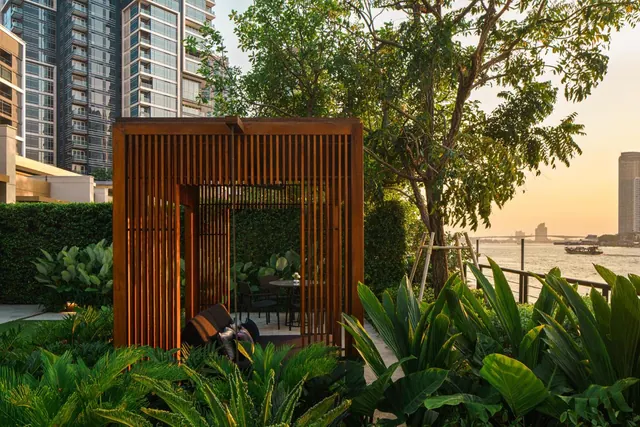 A slatted wooden pavilion with a small table and chairs stands among tropical plants in a garden, with tall towers and a river at sunset behind it.