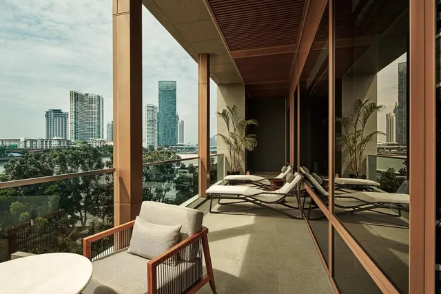A covered balcony with an armchair, a round marble table and lounge chairs overlooks high-rise towers and a river.