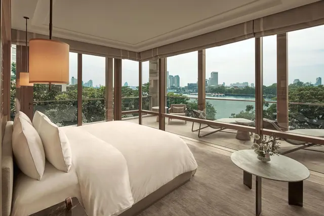 A bedroom with a king bed faces floor-to-ceiling windows that open onto a balcony with loungers, overlooking a river and city buildings.