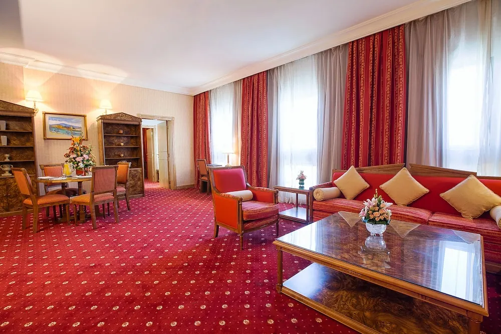 A furnished suite living area with a red sofa, armchairs, a glass-topped coffee table, a small dining table and tall curtained windows.