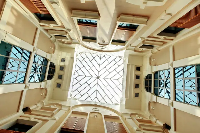 Looking up at a cream-coloured atrium with a geometric glass skylight and tall windows along both sides.