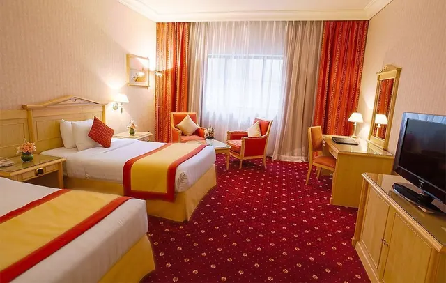 A twin-bedded hotel room with a red patterned carpet, two armchairs by a window with sheer curtains, a work desk, and a flat-screen television on a wooden cabinet.