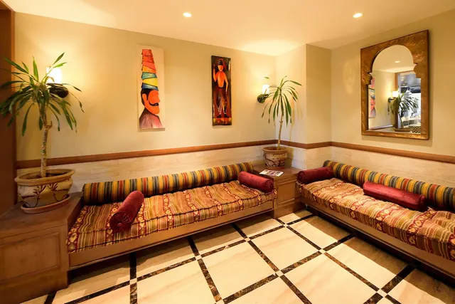 A seating area with built-in cushioned banquettes, potted palms, two wall paintings, a carved wall mirror and a marble tiled floor.
