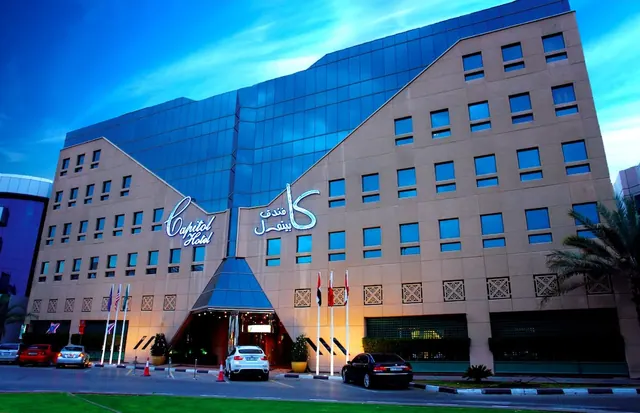 Exterior of the Capitol Hotel at dusk, with illuminated signage, flags, and cars parked at the entrance.