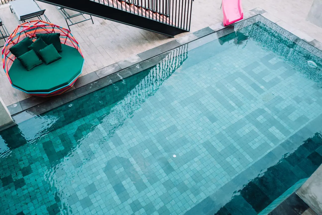 Elevated view of a mosaic-tiled swimming pool with a green circular daybed with cushions and grey sun loungers beside it, and a pink slide at the top edge of the frame.
