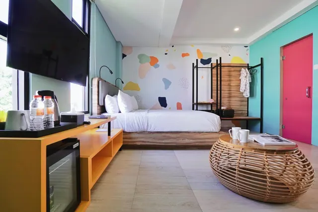A bedroom with a double bed against a multicolored painted wall, a wall-mounted television, a minibar, a kettle and a wicker side table.