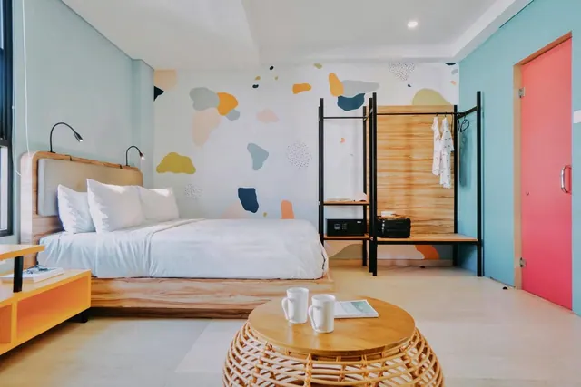 A bedroom with a king bed in front of a colourful abstract painted wall, an open clothing rack with a safe, and a round rattan table holding two mugs and a magazine.