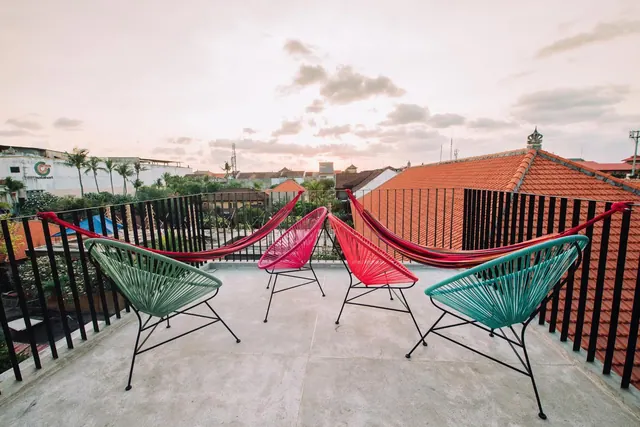 A rooftop terrace with four colorful woven chairs and two hammocks, with rooftops, palm trees and a pale sky visible beyond the railing at dusk.