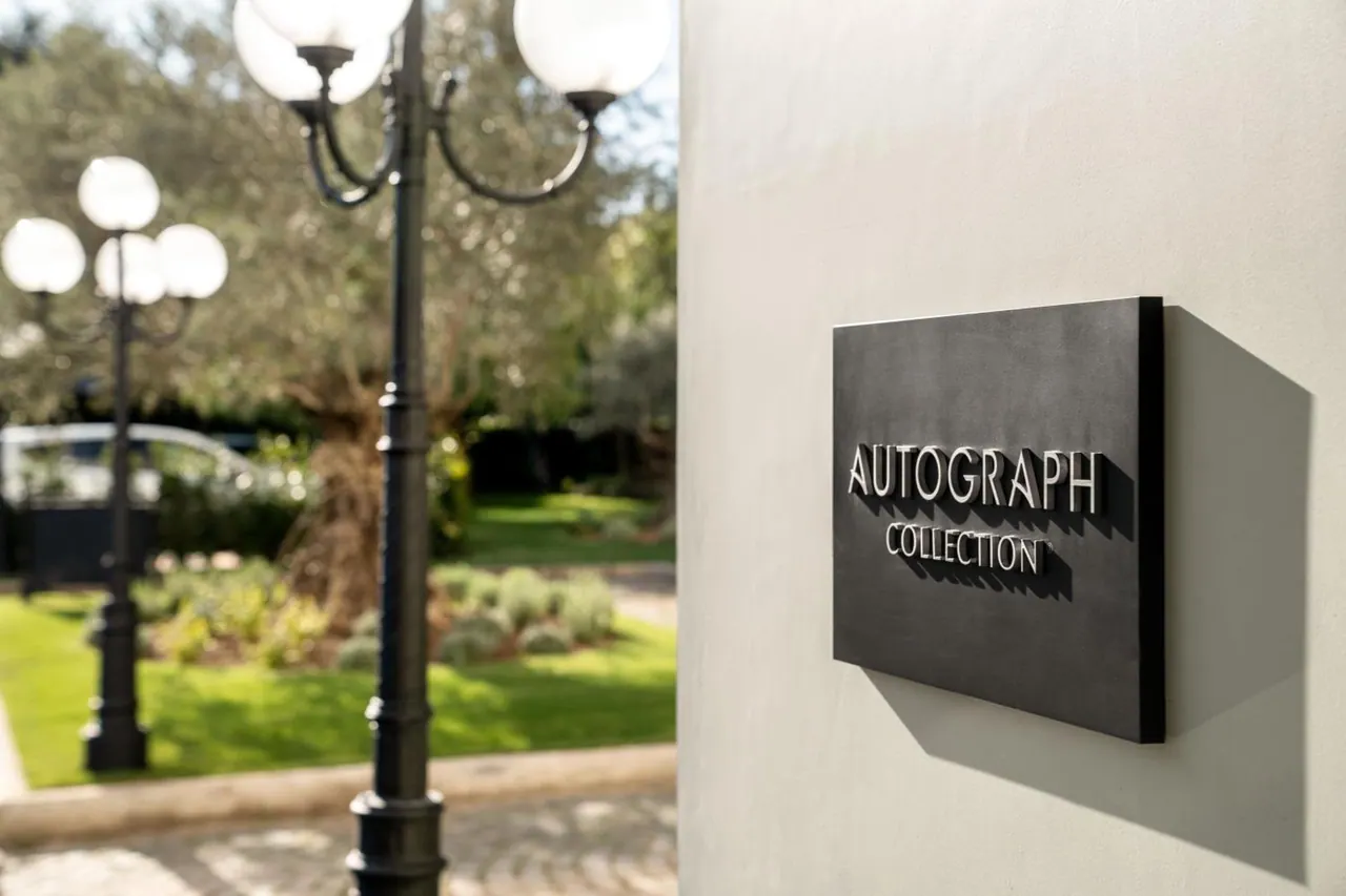 A dark metal sign reading AUTOGRAPH COLLECTION is mounted on a pale wall, with lamp posts and garden greenery blurred behind it.