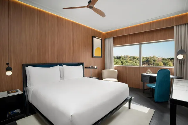 A bedroom with a made king bed, wood-panelled walls, a ceiling fan, a desk with a teal chair, and a window showing trees outside.