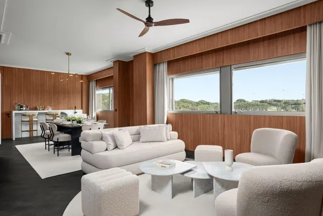 A living area with a cream sofa, armchairs and marble coffee tables, with a dining table and bar counter beyond, and windows looking out onto trees.