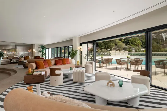 A lounge with peach sofas, cushions, a striped rug and white coffee tables, with floor-to-ceiling windows overlooking an outdoor pool and tables and chairs.