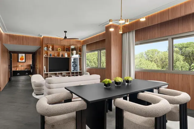 A staged open-plan living and dining room with wood-panelled walls, a black dining table with upholstered chairs, a sofa, a wall-mounted television and windows looking onto trees.