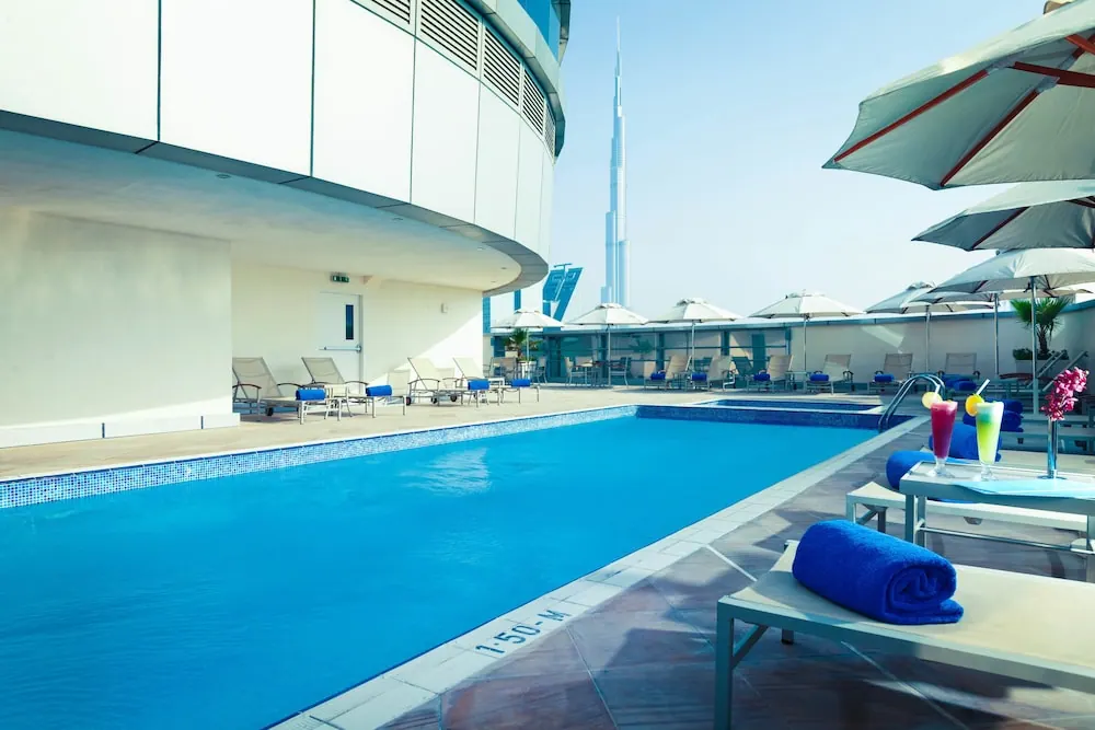 A rooftop swimming pool with blue towels, loungers and umbrellas, with the Burj Khalifa visible in the background.