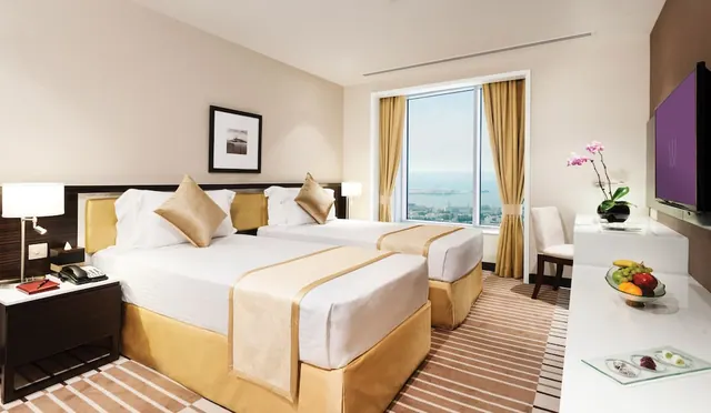 A bedroom with two single beds with gold and white bedding, a wall-mounted television, a desk, and a window with a view over water and city buildings.