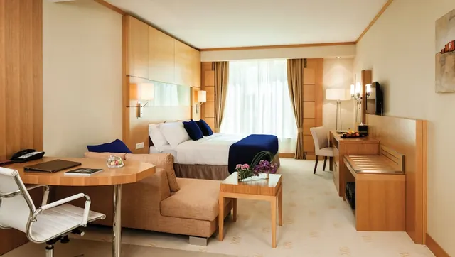 A hotel bedroom with a double bed under a navy throw, a sofa and armchair seating area, a round desk with a chair, and a wall-mounted television.