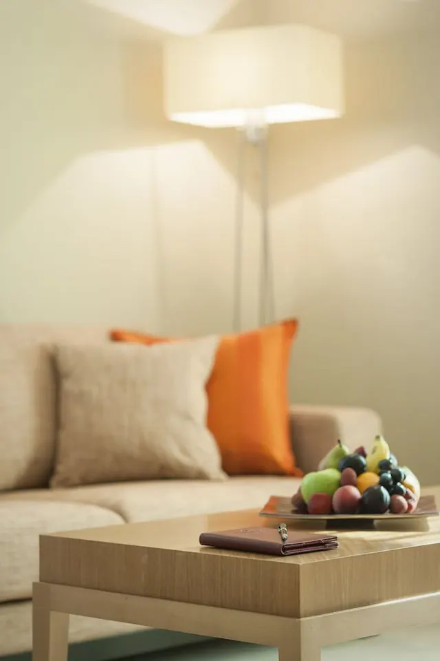 A living area with a beige sofa, beige and orange cushions, a lit floor lamp, and a wooden coffee table holding a fruit bowl and a leather folder.