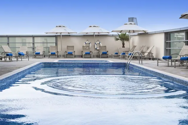 A rooftop swimming pool with blue-cushioned sun loungers and cream umbrellas along the surrounding deck under a clear blue sky.