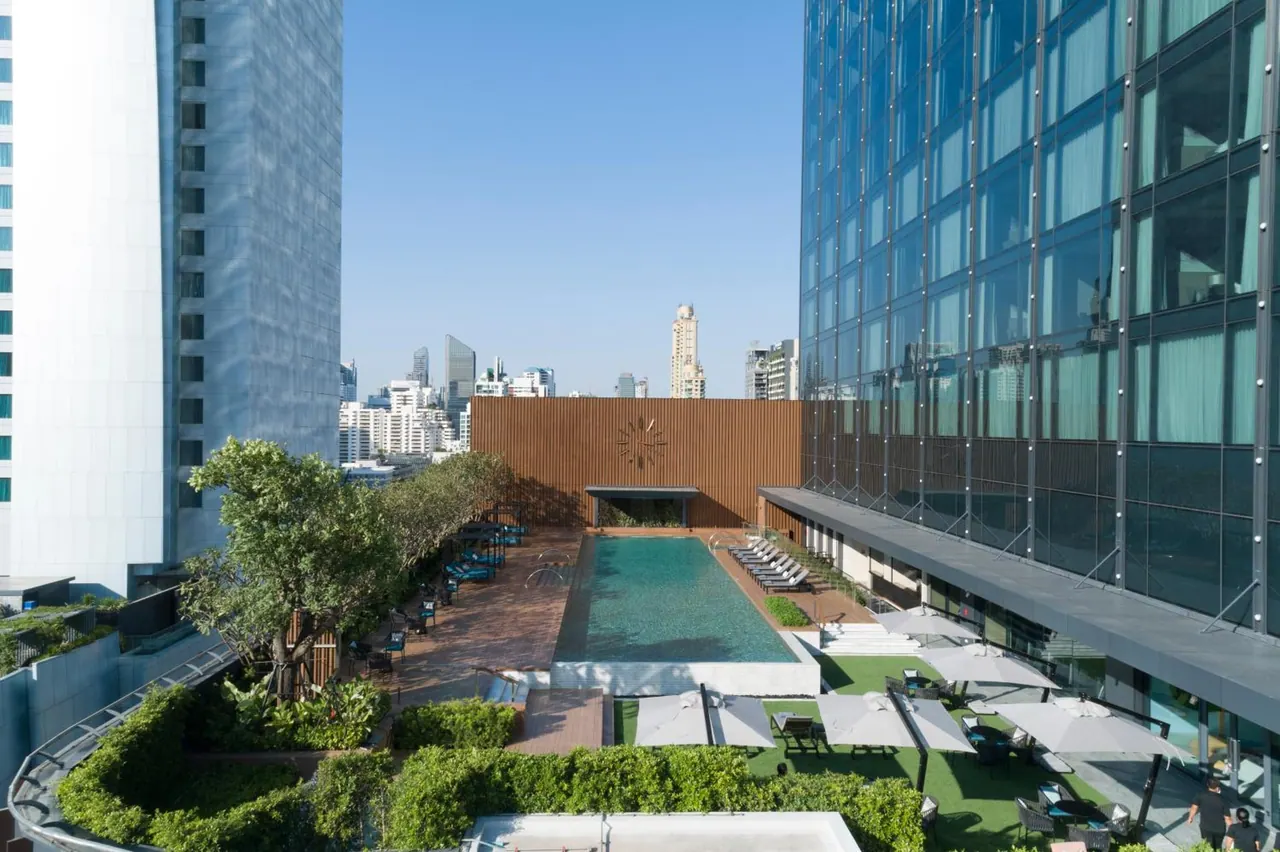 Elevated view of an outdoor swimming pool with sun loungers and grey umbrellas on the grass, flanked by glass towers, with a city skyline behind.