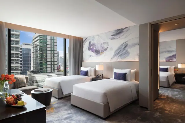 Two white twin beds stand in a bedroom with floor-to-ceiling windows looking out over high-rise buildings, with a sofa and a large abstract painting above the headboard.