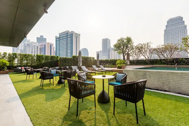 A rooftop terrace with artificial grass, dark woven chairs and a round table, beside a swimming pool with city towers beyond.