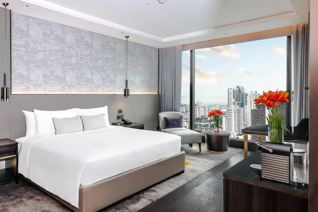 A king bed sits beside floor-to-ceiling windows overlooking a city skyline, with a coffee machine on a console in the foreground.