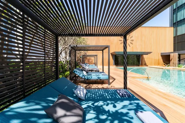 Blue daybeds sit under a slatted pergola beside a swimming pool on a wooden deck, with a timber-clad building behind.