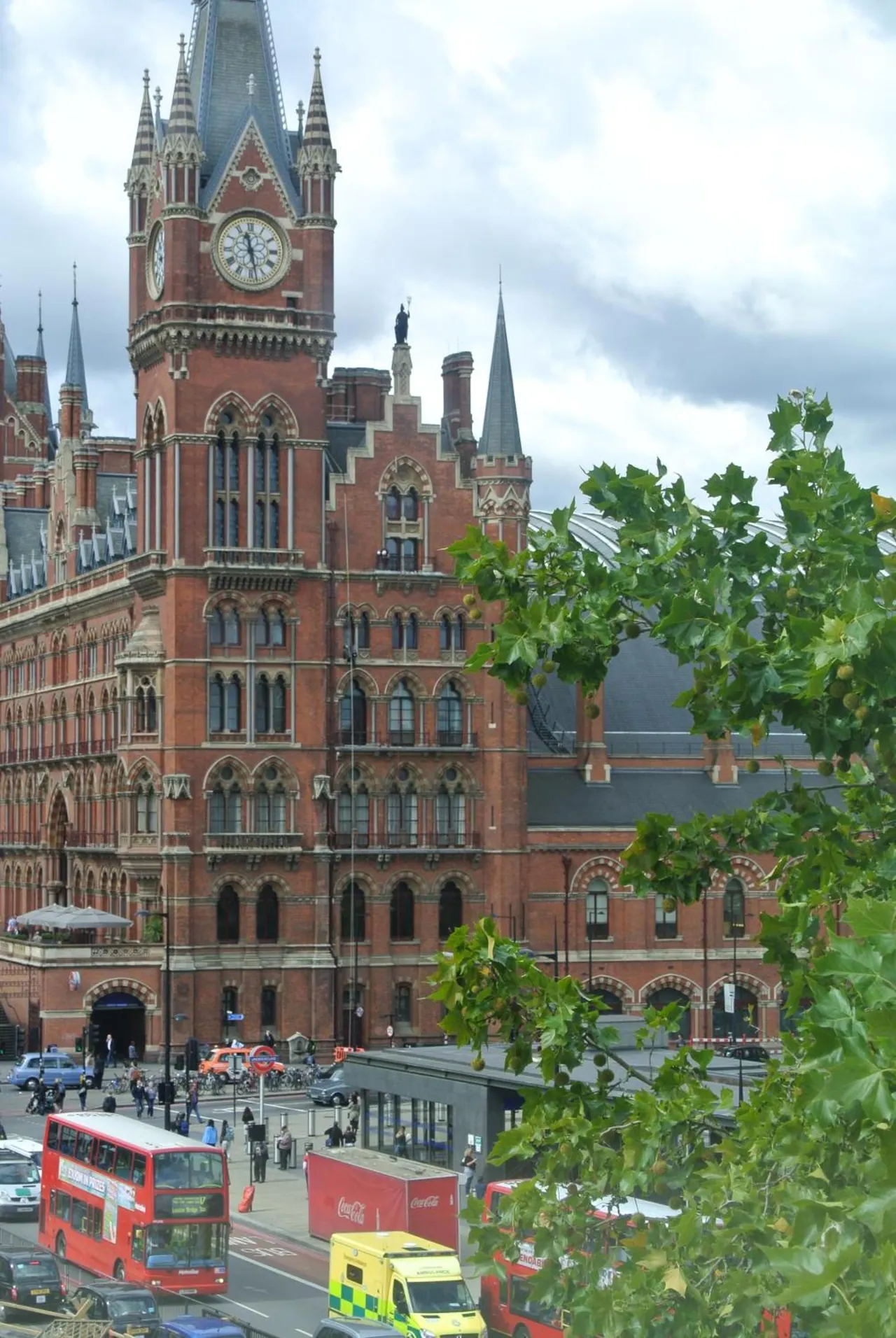 A view past green tree branches of the red brick Gothic clock tower of St Pancras station, with double-decker buses and an ambulance on the street below.