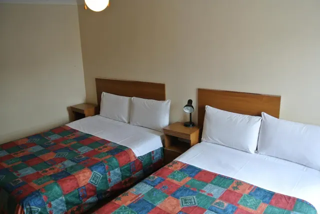 A bedroom with two double beds with white pillows and patchwork bedspreads, wooden headboards, and a small bedside lamp on a wooden nightstand.