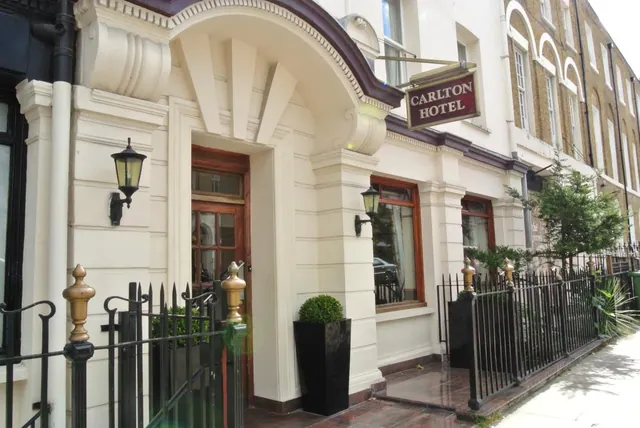 The entrance of the Carlton Hotel, with a cream facade, arched canopy, wooden door, wall lanterns and black iron railings.