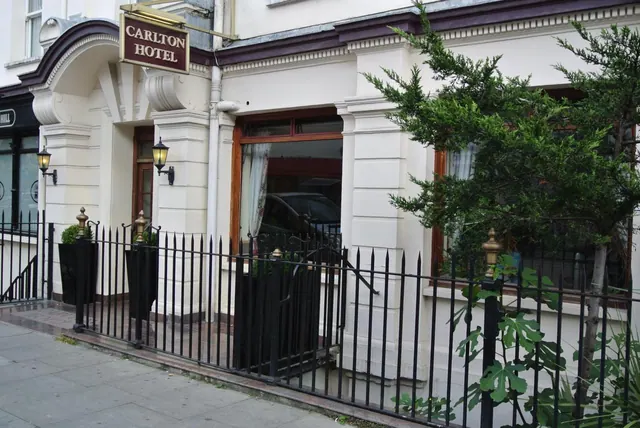 The facade of the Carlton Hotel is shown with its name sign above the entrance, black iron railings along the pavement and a tree to the right.