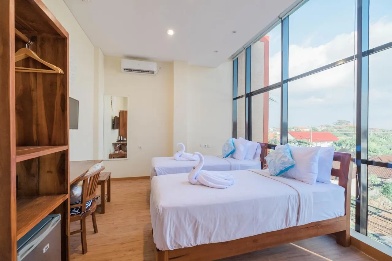 A hotel bedroom with two beds dressed in white linen and swan-shaped towels, a wooden wardrobe, a desk and chair, and floor-to-ceiling windows looking out over houses and trees.