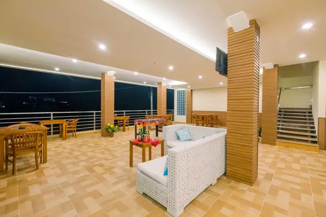 An open-sided terrace at night with wooden dining tables, white wicker sofas, a foosball table and a wall-mounted television.