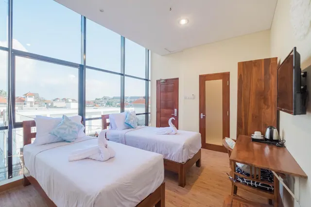 A bedroom with two single beds with white linen and towel swans, floor-to-ceiling windows, a wall-mounted TV, and a wooden desk with a kettle.
