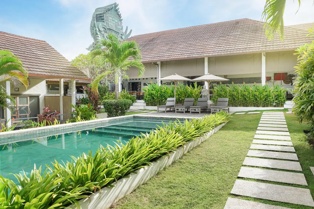 A rectangular swimming pool with a planted edge, sun loungers and umbrellas on the lawn, and a large green Garuda statue rising behind the building.
