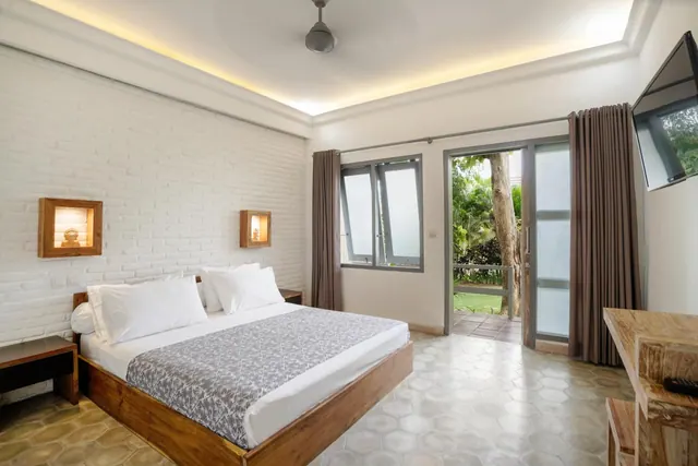 A bedroom with a king bed, a white brick wall with lit wall niches, a ceiling fan, a wall-mounted television, and a glass door opening onto a garden with trees.