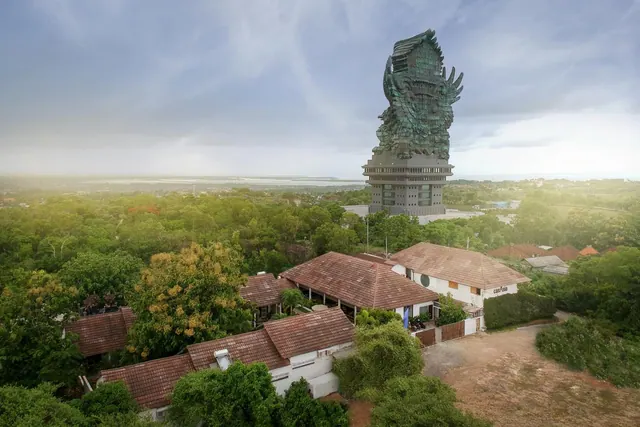 Elevated view over tree-covered land and tiled-roof buildings toward the Garuda Wisnu Kencana statue under a hazy sky.
