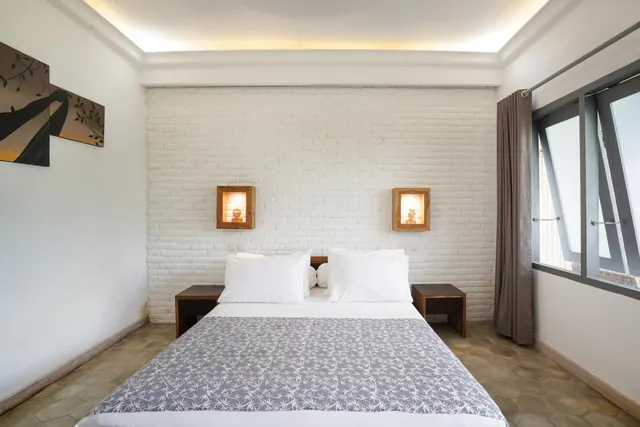 A double bed with white pillows and a grey patterned throw sits in front of a whitewashed brick wall with two lit wall niches holding small statues.