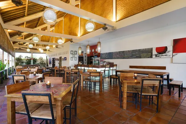 A dining room with wooden tables and chairs, woven pendant lamps, a chalkboard menu and a bar counter, with plants visible beyond the open side wall.